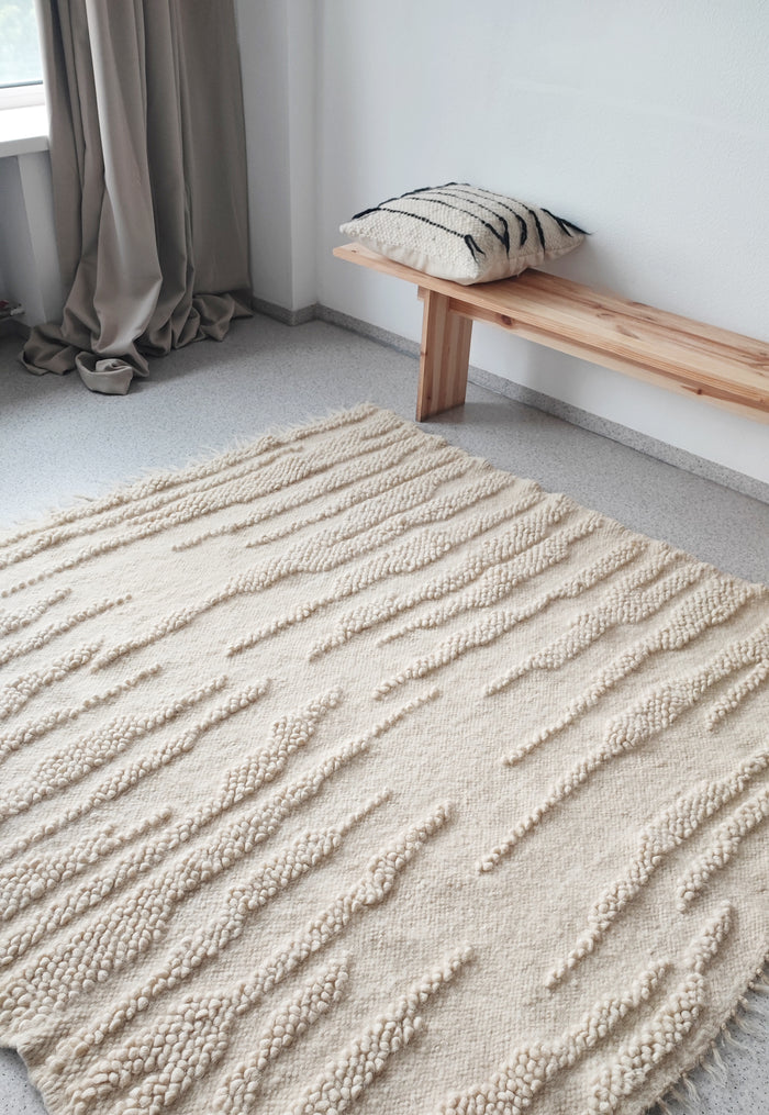 Textured beige rug on a gray floor with a wooden bench and striped pillow in the background.