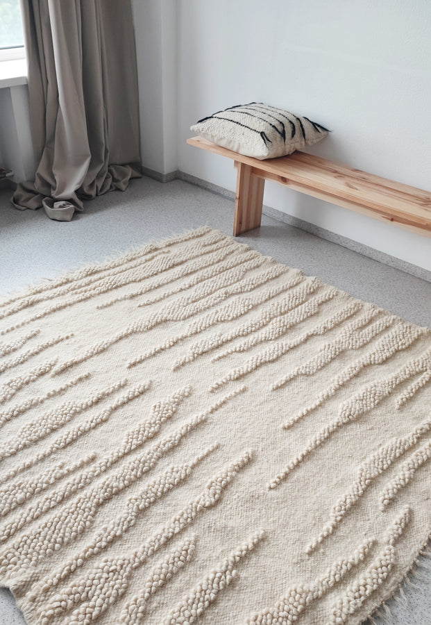 Textured beige rug on a gray floor with a wooden bench and striped pillow in the background.