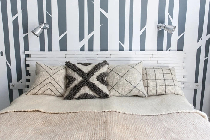 A styled bed with a white slatted headboard, textured cream bedding, and four decorative throw pillows in neutral tones, set against a wall with vertical gray and white tree branch patterns and two silver wall sconces.