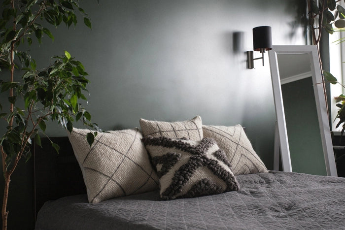 A styled bedroom scene featuring textured throw pillows on a bed, including a central black and cream patterned pillow, set against a dark gray wall with a wall lamp and a full-length mirror.