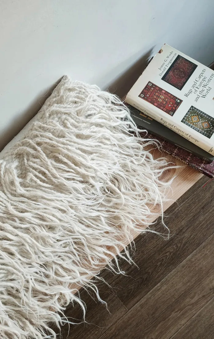 A cream-colored shaggy throw pillow with long fringe, styled on a wooden floor next to a book.