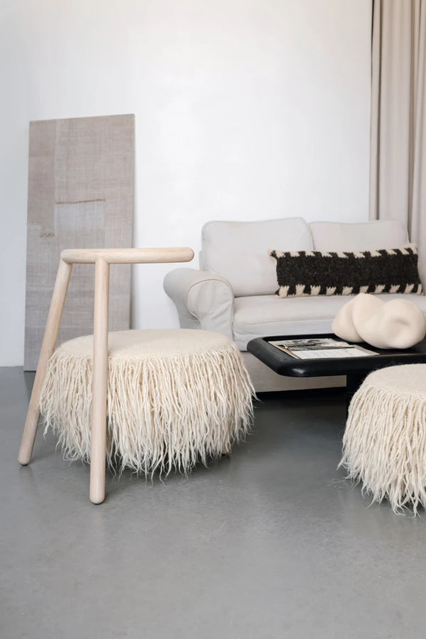 A modern living room scene featuring a light wood Mapico armchair with a cream shaggy fringe pouf seat, a matching cream pouf on the floor, a white sofa, and a black coffee table and mapico lumar pillow