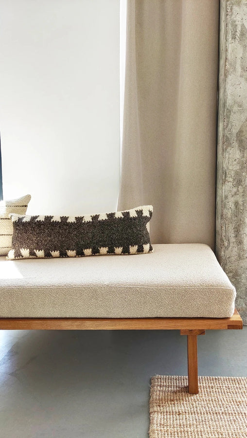 A modern daybed with a textured cream bouclé cushion and wooden frame, styled with a dark brown and cream patterned knit lumbar pillow in a minimalist room setting.