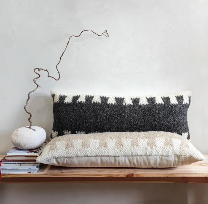 Two decorative throw pillows displayed on a wooden shelf against a textured light gray wall. The taller pillow is black and cream with a geometric pattern, and the shorter pillow in front is cream with a textured geometric design.