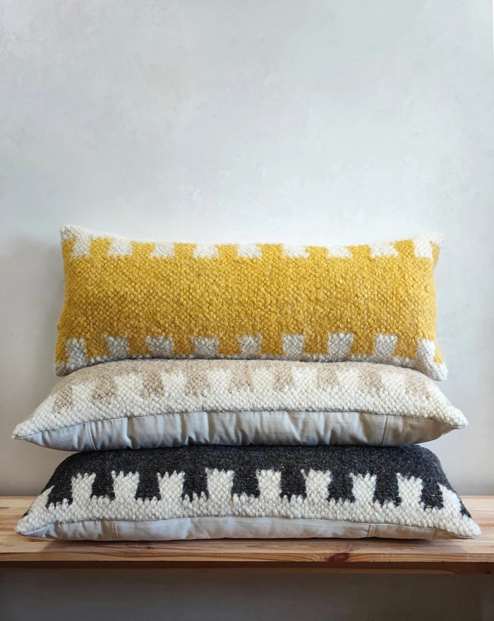Three rectangular woven throw pillows stacked vertically on a wooden shelf against a light textured wall. The top pillow is mustard yellow and off-white, the middle is cream and beige, and the bottom is dark gray and cream.