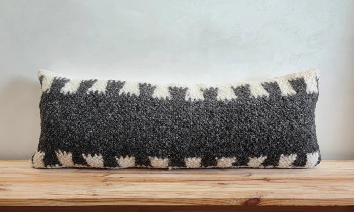 A rectangular lumbar throw pillow with a textured black and off-white geometric pattern, resting on a wooden surface against a neutral gray wall.
