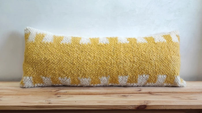A rectangular woven throw pillow with a yellow and white geometric pattern, resting on a light wooden surface against a textured off-white wall.