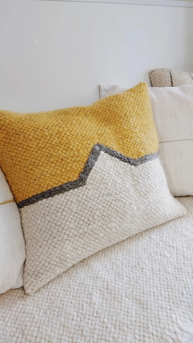 A textured woven throw pillow with a yellow top half and a cream bottom half, separated by a gray zigzag line, resting on a white textured blanket.