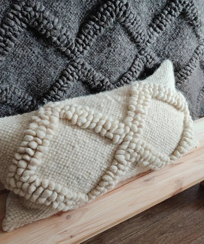 A close-up of a cream-colored woven throw pillow with chunky textured pom-pom trim, resting on a light wood surface in front of a dark gray knitted blanket.
