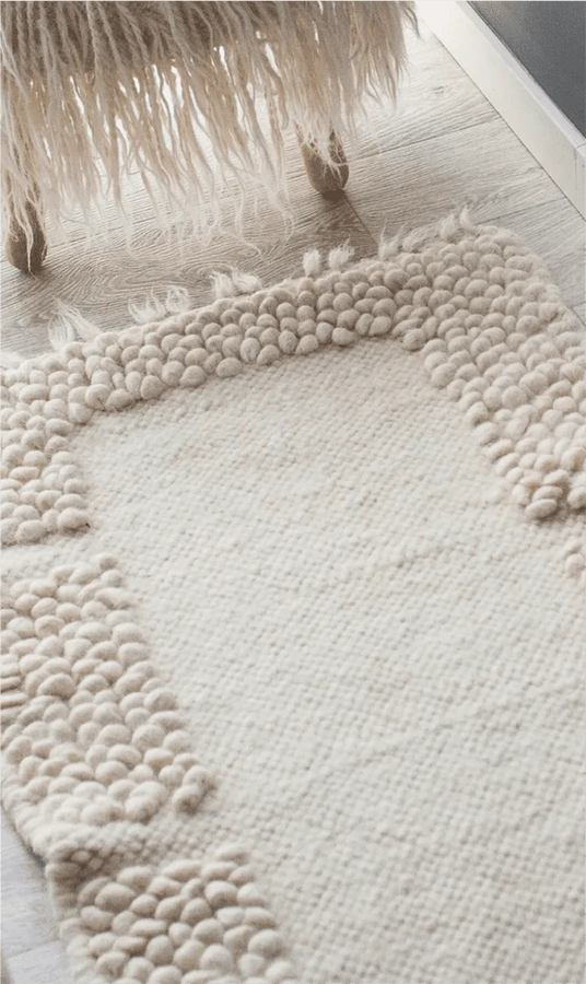 A close-up of a cream-colored textured rug with chunky looped borders, shown on a light wood floor with the fringe of a sheepskin or faux-shearling chair visible at the top. 